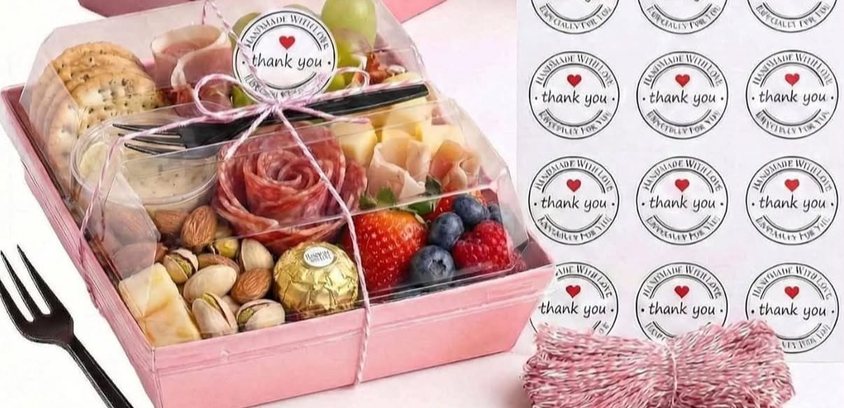 Pink square box with clear lid holding a charcuterie and fruit selection, with thank-you stickers and twine